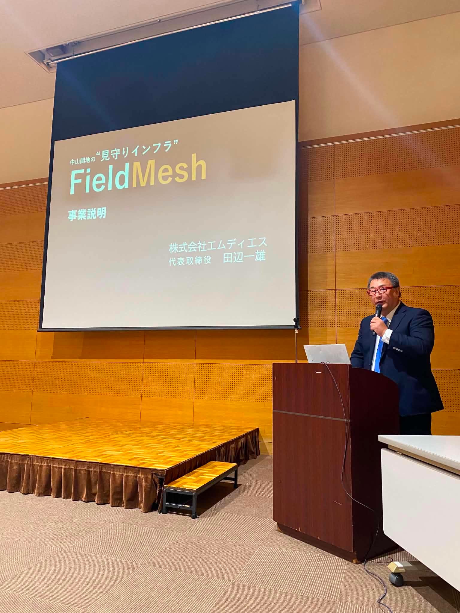 演台でマイクを持って話す田辺一雄。背後のスクリーンに「中山間地の“見守りインフラ” FieldMesh 事業説明 株式会社エムディエス 代表取締役 田辺一雄」と映っている。